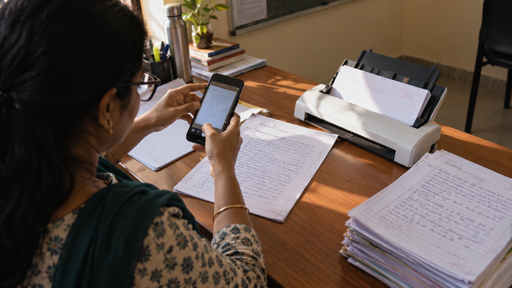 A staff member photographs a handwritten answer sheet on a phone, with a desktop scanner and remaining papers on the desk